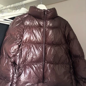 Brun pufferjacka med hög krage - Snygg pufferjacka i mörkbrunt med glansig finish och hög krage. Jackan har quiltad design, dragkedja framtill och praktiska fickor med dragkedja. Perfekt för kalla dagar och har en cool, oversized look som är trendig just nu. Ny pris runt 1200