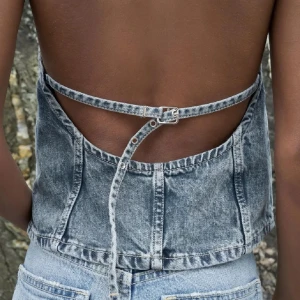 Grå jeanslinnetopp med öppen rygg - Säljer en snygg grå jeanslinnetopp med öppen rygg och justerbar snörning bak. Toppen har en rak passform och är ärmlös med hög halsringning. Perfekt för dig som vill sticka ut med en cool denimlook i sommar.