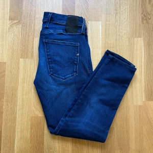 Replay Anbass Hyperflex jeans blå - Snygga Replay Anbass Hyperflex jeans i mörkblå tvätt med klassisk femficksdesign. Jeansen har raka ben och normal passform, tillverkade i stretchigt jeansmaterial för extra komfort. Perfekta för dig som gillar stilrena och bekväma jeans.