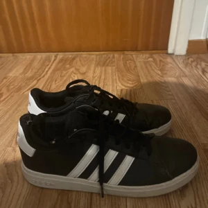 Svarta Adidas sneakers med vita ränder - Säljer ett par klassiska svarta Adidas sneakers med de ikoniska vita ränderna på sidorna och vit sula. Skorna har snörning och en låg profil som passar till många olika stilar. Perfekta för dig som gillar sportig och enkel design.