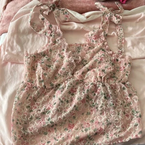 Blommig rosa linnetopp från Romwe - Supersöt rosa linnetopp från Romwe med broderade blommor i vitt och grönt. Toppen har knytbara axelband, spetsdetaljer och smockad rygg för extra komfort. Perfekt för dig som gillar romantisk och feminin stil!