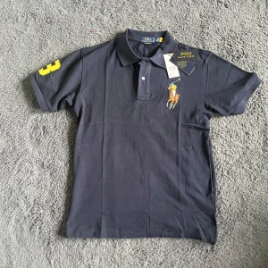 Ralph Lauren pike  - Helt ny Snygg mörkblå pike från Polo Ralph Lauren med klassisk polokrage och korta ärmar. Ikonisk broderad polospelare på bröstet och gul siffra 3 på ena ärmen. Tillverkad i mjuk bomull, perfekt för en chill och stilren look.  Axelbredd 47cm längd 71cm :) ny pris 125 dollar 