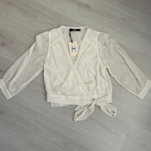 Vit prickig omlottblus från Bik Bok - Supersöt vit blus från Bik Bok i omlottmodell med knyt i midjan. Blusen har diskreta prickar, transparenta trekvartsärmar och dekorativ kant med små bollar längs v-ringningen. Perfekt för att ge din outfit en romantisk touch.