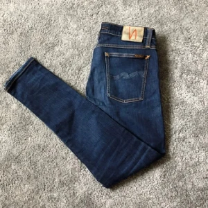 Blå skinny jeans från Nudie Jeans - Säljer ett par mörkblå skinny jeans från Nudie Jeans i storlek 32/32. Jeansen har klassiska fem fickor, orangea sömmar och snygga slitningar på benen. Materialet är mjukt jeans och passformen är tight med smal siluett. Kan skickas fler bilder annars.32/32 SLIM FIT