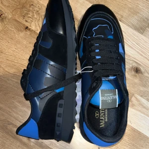 Valentino Garavani Rockrunner sneakers - Säljer ett par Valentino Garavani Rockrunner sneakers i svart och blått med coolt camouflagemönster. Skorna har snörning, platt sula och rund tå. Materialmix av skinn och syntet med detaljer i mocka. Perfekt för dig som gillar statement-sneakers med lyxig vibe. De är helt o använda och i helt nytt skick inga problem med dem 