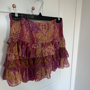 Paisleymönstrad volangkjol från Zara - Supersöt kort kjol från Zara med paisleymönster i lila, rosa och gult. Kjolen har flera lager volanger och smockad midja som ger en skön och snygg passform. Perfekt för dig som gillar boho-vibe och färgglada plagg.