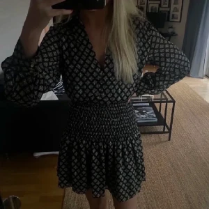 Svartvit kortklänning med mönster - Supersnygg kortklänning från Zara i svart och vitt med rutigt mönster. Klänningen har långärmade puffärmar, v-ringad hals och smockad midja som ger en cool siluett. Materialet är mjukt och lätt, perfekt för dig som gillar trendiga detaljer.