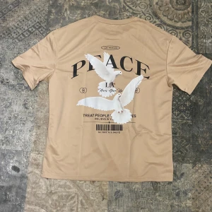 Beige PEACE t-shirt med duvor - Snygg beige t-shirt med tryck av två vita duvor och texten PEACE på ryggen. Framsidan har ett mindre tryck med duva och PEACE. Rund hals och korta ärmar, tillverkad i mjukt material som känns skönt mot huden. Perfekt för dig som gillar streetwear med budskap.