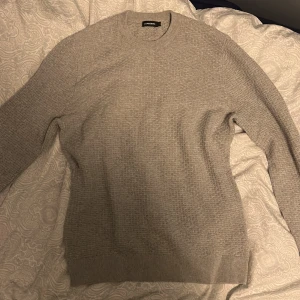Grå stickad tröja från J.Lindeberg - Snygg grå stickad tröja från J.Lindeberg i storlek M. Tröjan har ett diskret strukturerat mönster och rund hals. Ribbstickade muddar vid ärmslut och nederkant ger en clean look. Perfekt för dig som gillar stilrena och enkla plagg.
