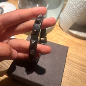 Valentino svart armband med nitar - Snyggt svart armband från Valentino Garavani med coola pyramidformade silverfärgade nitar runt om. Gjort i äkta skinn och har ett justerbart spänne. Perfekt accessoar för dig som vill ha en edgy och stilren look. Allt OG på bilden tillkommer. Defekter syns på bild(lädret har gått upp, men borde gå att limma)🌟