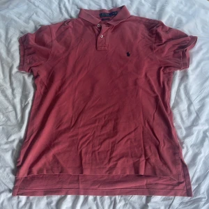 Rosa Polo Ralph Lauren t-shirt L - Snygg rosa t-shirt från Polo Ralph Lauren i storlek L. Klassisk pikémodell med krage, två knappar och den ikoniska broderade loggan på bröstet. Tillverkad i 100% bomull för skön och luftig känsla. Custom fit som sitter riktigt bra. Den har en pytteliten vit fläck nära loggan. Priset kan diskuteras. 