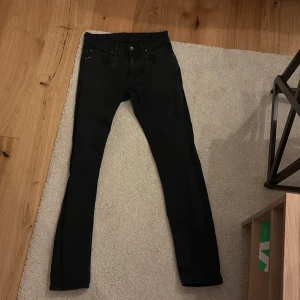 Svarta Tiger of Sweden jeans - Svarta jeans tillverkade i Italien. Modellen har klassisk femficksdesign, raka ben och normal passform. Jeansen är i svart denim med svarta sömmar och har bälteshällor samt knappstängning framtill. Perfekt för dig som gillar stilrena och enkla jeans.