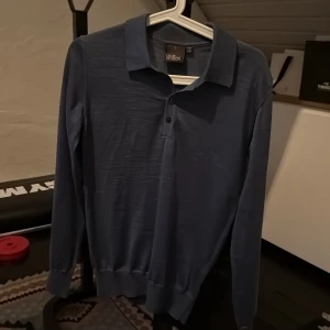 Blå pikétröja från Oscar Jacobson - Säljer en stilren blå pikétröja från Oscar Jacobson. Tröjan har klassisk krage, knäppning framtill och långa ärmar. Tillverkad i Italien och passar perfekt för en dressad men avslappnad look.