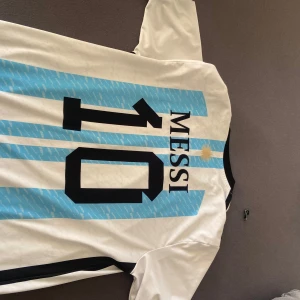 Argentina Messi 10 fotbollströja - Säljer en klassisk Argentina fotbollströja med Messi och nummer 10 på ryggen. Tröjan är vit med ljusblå ränder, svarta detaljer och guldiga emblem. Perfekt för dig som älskar fotboll och vill stötta landslaget.