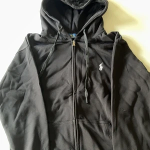 Svart hoodie med dragkedja från Polo Ralph Lauren - Svart hoodie från Polo Ralph Lauren med dragkedja framtill och vit broderad logga på bröstet. Tröjan har huva med snörning och långa ärmar. Perfekt för en avslappnad stil. Helt ny, oanvänd