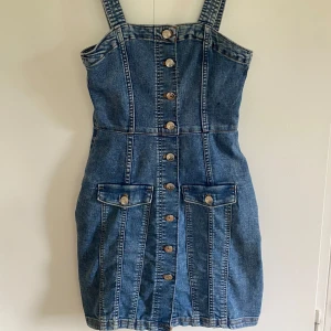 Jeansklänning med knappar från H&M Divided - Säljer en jeansklänning från H&M Divided i klassisk blå denim. Klänningen har breda axelband, knappar framtill och dekorativa fickor med knappdetaljer. Perfekt för en trendig och avslappnad look.