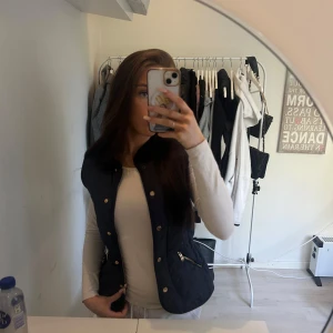 Mörkblå quiltad väst med dragkedjor - Snygg mörkblå quiltad väst med ståkrage och guldiga knappar framtill. Två snedställda fickor med dragkedja och justerbar midja baktill. Perfekt att styla över en långärmad tröja för en trendig look. Aldrig använd med prislapp kvar. 