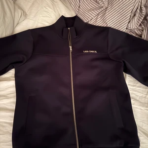 Svart jacka/tröja från Les Deux med dragkedja - Säljer en svart jacka från Les Deux med hög krage och silverfärgad dragkedja. Jackan har två fickor framtill och stilren logga på bröstet. Perfekt för dig som gillar enkel och snygg design. Tröjan köptes på kidsbrandstore i köpcentret väla. Skick 9/10