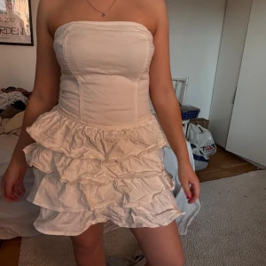 Vit volangklänning med axelbandslös design - Säljer en söt axelbandslös vit klänning med flera volanglager och figurnära passform upptill. Klänningen är kort och har en romantisk känsla, perfekt för sommarens tillfällen. Materialet känns mjukt och lätt.