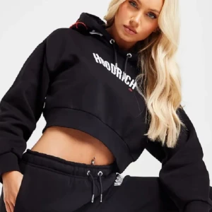 Svart croppad hoodie från Hoodrich - Säljer en svart croppad hoodie från Hoodrich med vit logga på bröstet och tryck på huvan. Tröjan har långa ärmar, huva med dragsko och en avslappnad passform. Perfekt för en cool och sportig stil.