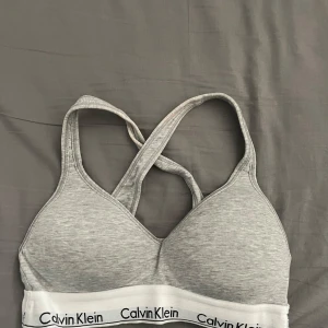 Grå bh från Calvin Klein - Bh i ljusgrått från Calvin Klein med klassisk logotyp på det vita resårbandet. Bh:n har korsade axelband i ryggen och är tillverkad i mjukt bomullsmaterial. Tyvärr är det lite hårfärg på banden men inte något man lägger märke till🥰