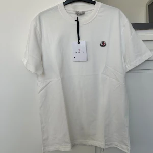 Vit t-shirt från Moncler - Säljer en klassisk vit t-shirt från Moncler med rund halsringning och Moncler-logga broderad på bröstet. Perfekt basic-plagg med stilren design och mjukt material.