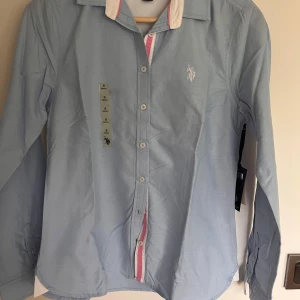 Ljusblå skjorta från U.S. Polo Assn - Säljer en ljusblå skjorta från U.S. Polo Assn med vita och rosa detaljer vid krage och knappslå. 
