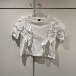Vit volangtopp från Gina Tricot - T-shirt med volanger på båda sidorna både fram och bak. Är lite kortare i modellen.  Strl 38 Använd fåtal gånger. Är i mycket fint skick.
