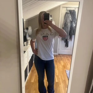 Vit t-shirt från Gant med logga - Vit t-shirt från Gant med stor logga och sköld på bröstet. Klassisk passform och rund halsringning. Perfekt till jeans eller shorts. 