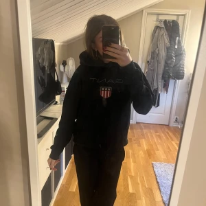 Svart hoodie från Gant med logga - Svart hoodie från Gant med broderad logga och sköld på bröstet. Klassisk huva och känguruficka framtill. 