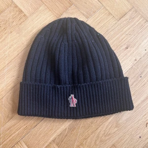 Svart ribbstickad mössa från Moncler - Svart ribbstickad mössa från Moncler med uppvikt kant och liten broderad logotyp framtill. Klassisk och stilren design som passar perfekt till kyliga dagar.
