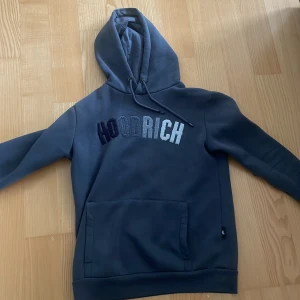 Mörkgrå hoodie från Hoodrich - Säljer en mörkgrå hoodie från Hoodrich i storlek XS men som också passar S Tröjan har broderad logga i olika grå nyanser på bröstet, huva med dragsko och en stor magficka. Perfekt för dig som   vill ha något bekvämt och snyggt.
