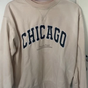 Beige sweatshirt med CHICAGO-tryck från H&M - Säljer en beige sweatshirt från H&M med stort CHICAGO-tryck i svart på bröstet. Tröjan har rund halsringning och långa ärmar. Perfekt för en avslappnad stil och enkel att matcha med jeans eller leggings.
