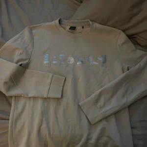 Beige sweatshirt från BOSS - Säljer en beige sweatshirt från BOSS med rund halsringning och tryckt logga i silver på bröstet. Tröjan har långa ärmar och ribbade muddar vid ärmslut och nederkant. Perfekt för en avslappnad stil.