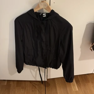 Svart vindjacka med huva - Säljer en svart vindjacka med huva och dragsko i midjan. Jackan har långa ärmar, dragkedja och knappar framtill samt elastiska muddar vid ärmslut. Perfekt för blåsiga dagar och enkel att matcha med olika outfits.❤️❤️ i storlek xs❤️