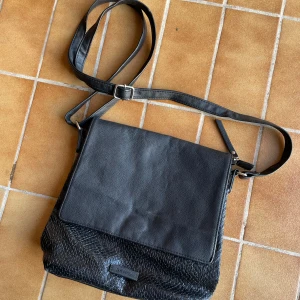 Svart axelväska från Don Donna - Säljer en svart axelväska från Don Donna med justerbar rem. Väskan har en slät ovandel och en flätad struktur på nedre delen, vilket ger en snygg kontrast. Perfekt för att bära dina viktigaste saker i vardagen.