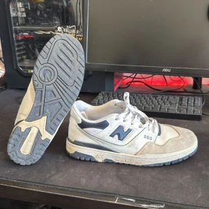 New Balance 550 sneakers i grått och vitt - Säljer ett par New Balance 550 sneakers i grått och vitt med mörkblå detaljer. Skorna har snörning, rund tå och platt sula. Perfekta för dig som gillar klassisk streetwear-stil och vill ha bekväma sneakers till vardags.