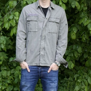 Grå overshirt med fickor - Säljer en stilren grå overshirt med två bröstfickor och klassisk krage. Jackan har lång ärm och stängs med dolda knappar framtill. Perfekt för lager-på-lager och enkel att matcha med jeans eller chinos. Märke black squad . Lappen saknas på ärmen men så det är bara knappar