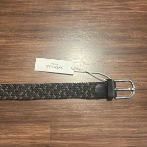 Grått flätat bälte från ONEWear Sweden - Snyggt grått flätat bälte från ONEWear Sweden med silverfärgat spänne och svarta skinn-detaljer vid ändarna. Perfekt accessoar för att lyfta din outfit med en stilren touch.