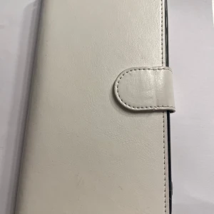 Vit plånboksväska för iPhone 8 Plus  - Säljer en stilren vit plånboksväska för mobil med plats för kort och ID-fönster. Väskan har magnetlås och är tillverkad i syntetmaterial. Perfekt för dig som vill ha allt samlat på ett ställe.