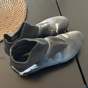 Svarta fotbollsskor från Puma - Säljer ett par svarta fotbollsskor från Puma med silvriga detaljer och logga på sidan. Skorna har en strumpliknande passform utan snörning och är tillverkade i syntetmaterial för bra grepp och komfort på planen.