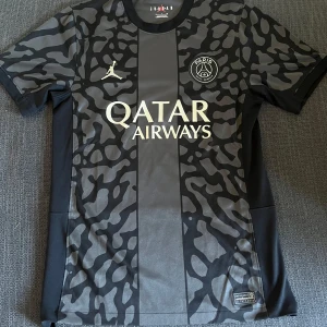 PSG matchtröja - Säljer en svart och grå PSG fotbollströja från Jordan med camoinspirerat mönster. Tröjan har korta ärmar, Paris Saint-Germain-logga på bröstet och Qatar Airways som sponsortryck. Perfekt för dig som gillar snygga och unika matchtröjor. Tröjan är 100% äkta. Tröjan är använd några gånger men är fortfarande i ett super bra skick. Skick: 9/10.