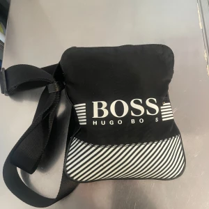 Svart axelväska från Hugo Boss - Snygg svart axelväska från Hugo Boss med vit logga framtill och diagonalrandigt mönster i vitt och svart på nedre delen. Väskan har justerbar axelrem och stängs med dragkedja. Perfekt för att bära det viktigaste. Finns i Göteborg och helst vill träffas! Öppet för byte 