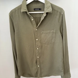 Olivgrön skjorta från J.Lindeberg - Säljer en olivgrön skjorta från J.Lindeberg i storlek M. Skjortan har klassisk krage, bröstficka och knappar framtill. Tillverkad i mjukt bomullstyg och har en diskret broderad logga vid nederkanten. Perfekt för en stilren look.