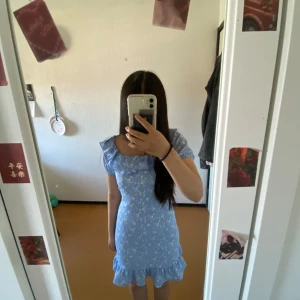 Blå blommig klänning från Gina Tricot - Supersöt blå klänning från Gina Tricot med vita blommor och volangdetaljer. Klänningen har korta ärmar, smockad rygg och volang längst ner. Perfekt för sommarens alla tillfällen!