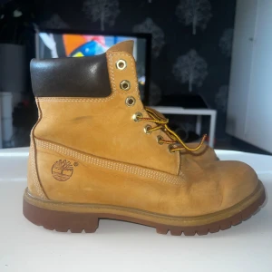Beiga Timberland boots 6 inch - Klassiska Timberland boots i beige nubuck med brunt vadderat skaft och guldfärgade metalldetaljer. Skorna har snörning och grov platt sula för extra grepp. Perfekta för höst och vinter. Endast lite sliten på toeboxen och några creases annars 10/10