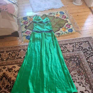 Grön långklänning i satin - Säljer en smaragdgrön långklänning i glansigt satinmaterial med hjärtformad urringning och markerad midja. Klänningen har axelbandslös design och en elegant, svepande kjol. Perfekt för bal eller fest.