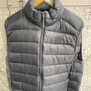 Grå dunväst från Lindbergh - Snygg grå dunväst från Lindbergh med quiltad design och hög krage. Västen har dragkedja framtill och en diskret logotyp på bröstet. Perfekt att bära över en tröja för extra värme under kyliga dagar. Säljer för att den är för stor för mig.