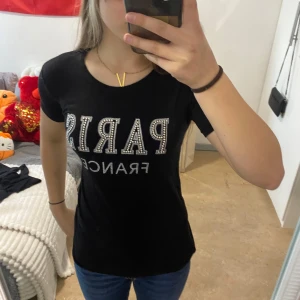 Svart t-shirt med strasstext och meshdetalj 50/st - Säljer en svart t-shirt med glittrande strasstexten 'PARIS FRANCE' framtill och dekorativa stenar på baksidan. T-shirten har korta ärmar och meshparti upptill vid axlarna. Perfekt för dig som gillar statement-plagg med lite extra bling.