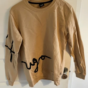 Beige sweatshirt från BOSS  - Säljer en beige sweatshirt från BOSS Orange med svart tryck längs ena ärmen och framtill. Tröjan har rund halsringning och långa ärmar. Perfekt för en avslappnad och stilren look.